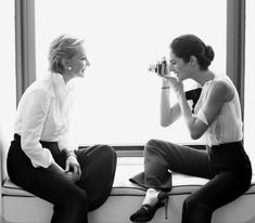 two women are sitting on a window sill and one is holding a camera