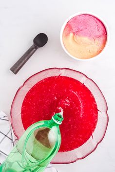 a red and green drink in a glass bottle next to a white plate with spoons