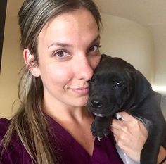 a woman holding a black puppy in her arms and smiling at the camera while wearing a purple shirt