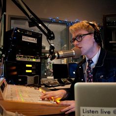 a man sitting in front of a microphone with headphones on and recording equipment behind him