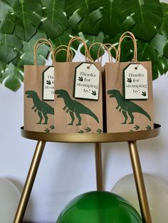 two brown bags with green dinosaurs on them sitting on a table next to balloons and a potted plant