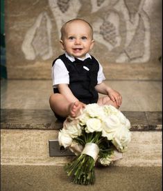 a baby boy sitting on the ground next to a bouquet of white flowers and smiling