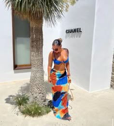 a woman in a multicolored dress is standing next to a palm tree and holding a handbag