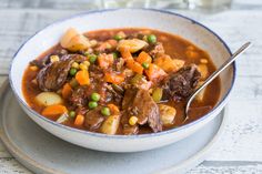 a bowl of beef stew with carrots, peas and potatoes on a white plate