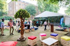 many people are standing around in the grass near some trees and benches with colorful cushions on them