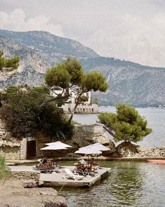 some people are sitting at an outdoor table by the water with umbrellas and chairs