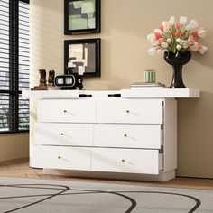 a white dresser with flowers on top in a room next to a window and framed pictures