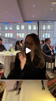 a woman sitting at a table drinking from a wine glass with people in the background