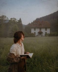 a woman standing in the middle of a field holding a book and looking at a house