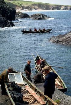 several people in canoes on the shore with rocks and water behind them, one man is holding something