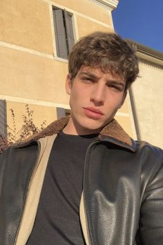 a young man wearing a leather jacket standing in front of a building and looking at the camera