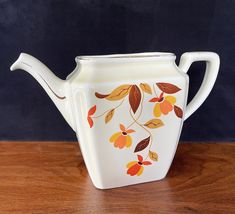 a white tea pot with orange and yellow leaves painted on the side sitting on a wooden table