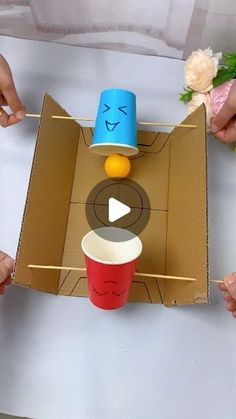 three people holding cups with faces painted on them in a cardboard box and one is opening the lid