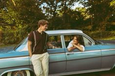 a man and woman leaning on the door of an old car in front of trees