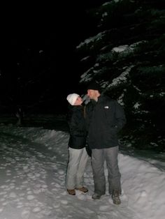 two people standing in the snow at night with their noses close to each other and trees behind them