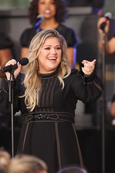 a woman standing in front of microphones on stage with other people behind her and singing