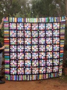 a man holding up a large quilt made from strips of multicolored fabric in the woods