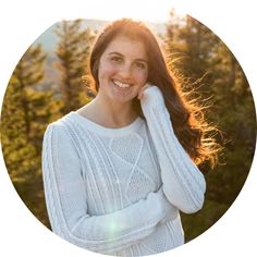 a woman with long hair wearing a white sweater smiles at the camera in front of trees