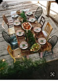 an outdoor dining table set with plates and bowls on it, surrounded by greenery