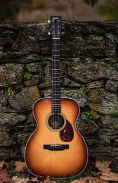 an acoustic guitar sitting on the ground next to a stone wall with grass and leaves around it