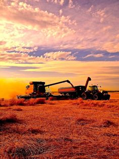 two large machines are in the middle of a wheat field as the sun goes down