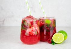 two glasses filled with liquid next to limes and strawberries on a white surface