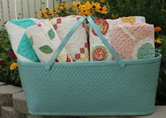 a blue basket filled with lots of different colored pillows next to some flowers and bushes