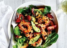 a salad with shrimp, avocado and spinach on a plate next to a glass of wine