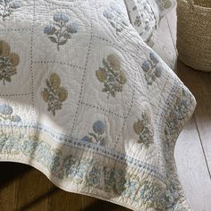 a white quilted bed with blue flowers on it and a basket next to it