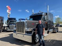 a man standing next to a large semi truck in a parking lot with other semi trucks behind him