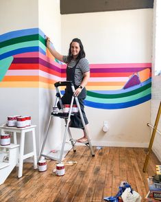 a woman standing on a ladder painting a wall with colorful stripes and rainbows in the background