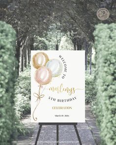 a sign with balloons on it in the middle of a path that is surrounded by trees