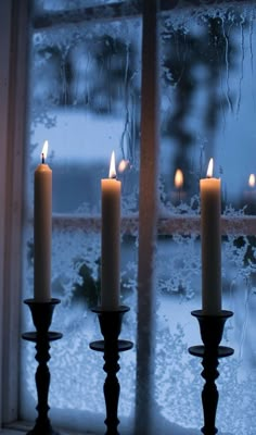 three lit candles sitting on top of a window sill in front of a snow covered window