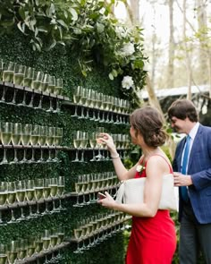 a man and woman standing next to each other in front of a green wall filled with glasses