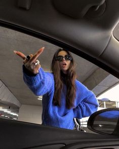 a woman in blue sweater and sunglasses holding up her hand while standing next to a car