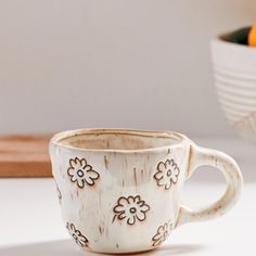 a white coffee cup sitting on top of a table next to a bowl filled with oranges