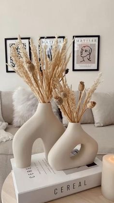 two white vases with dried flowers in them on a table next to a candle