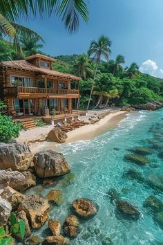 a house on the beach with clear blue water and rocks in front of it, surrounded by palm trees