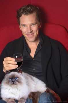 a man sitting in a red chair holding a glass of wine with a cat on his lap