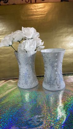 two white vases with flowers in them sitting on a shiny tableclothed surface