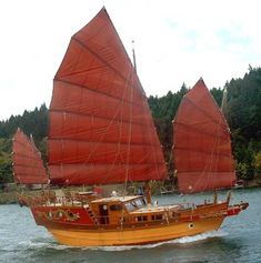 an orange sailboat with red sails on the water in front of trees and hills