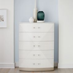 a white chest of drawers sitting next to a vase on top of a wooden floor