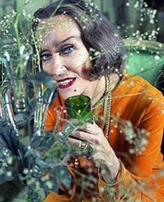 a woman in an orange dress is holding a vase with flowers on it and looking at the camera