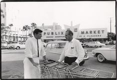 two men standing next to each other in front of a car with shopping carts on it