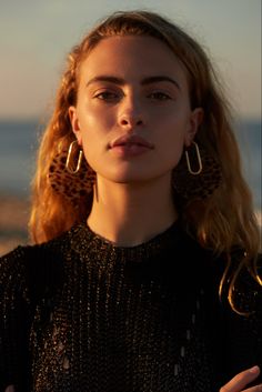 a woman with long blonde hair wearing large hoop earrings and black top standing on the beach