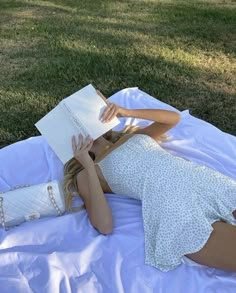 a woman is laying on the grass reading a book and holding a white purse in her hand