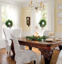 a dining room table with white chairs and wreaths on the wall behind it in front of two windows