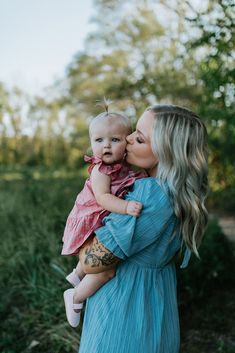 a woman holding a baby in her arms and kissing it's face with trees in the background