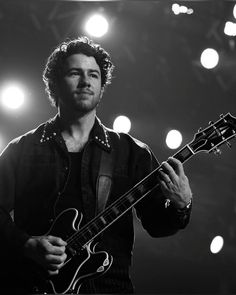 a man holding a guitar in his right hand and looking at the camera with lights behind him