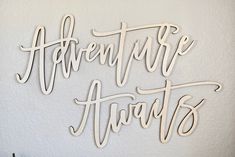 the word adventure awaits written in cursive type on a white wall next to a potted plant